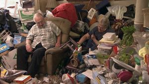 An older couple sitting among piles of stuff in a hoarder house